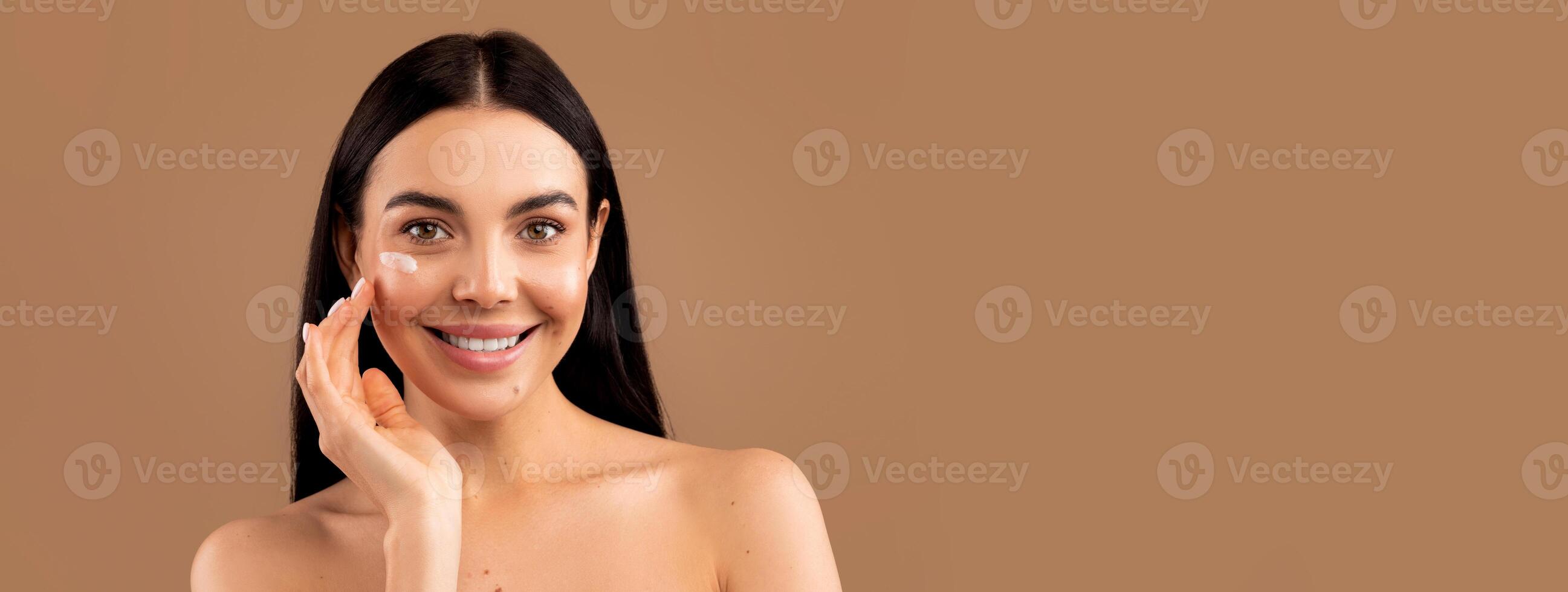 Cheerful sensual naked long-haired brunette millennial woman with smile on her face using facial eye cream for young smooth glowing skin, isolated on beige studio background, copy space photo
