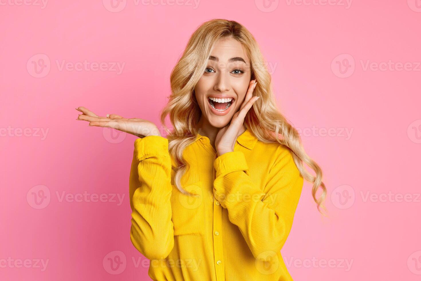 A young woman with blonde hair and a bright smile is wearing a yellow button-up shirt and posing with her hand open and palm facing outward. The background is a solid pink color. photo