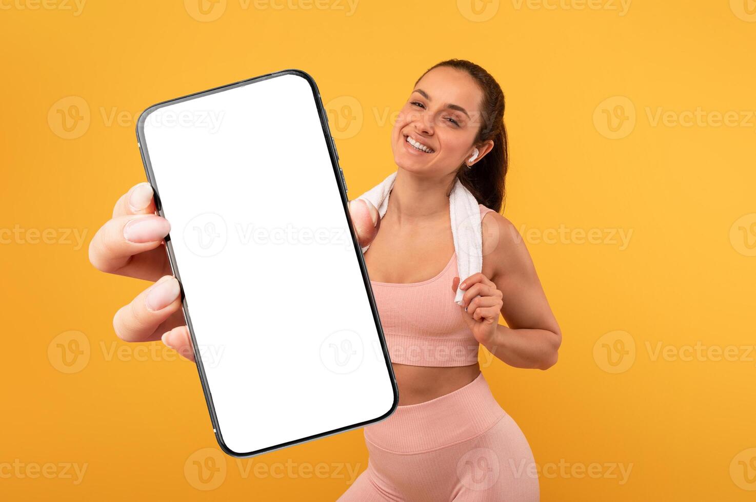 A young woman smiles broadly while holding a blank smartphone in one hand and a towel draped over her shoulder. She is dressed in athletic gear and stands in front of a vibrant yellow wall. photo