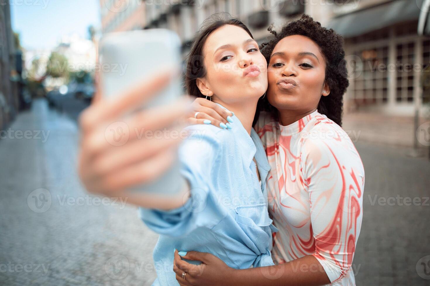 Two young multiethnic women taking a selfie on a city street. They are both smiling and looking at the camera. The woman with long brown hair is holding the phone photo