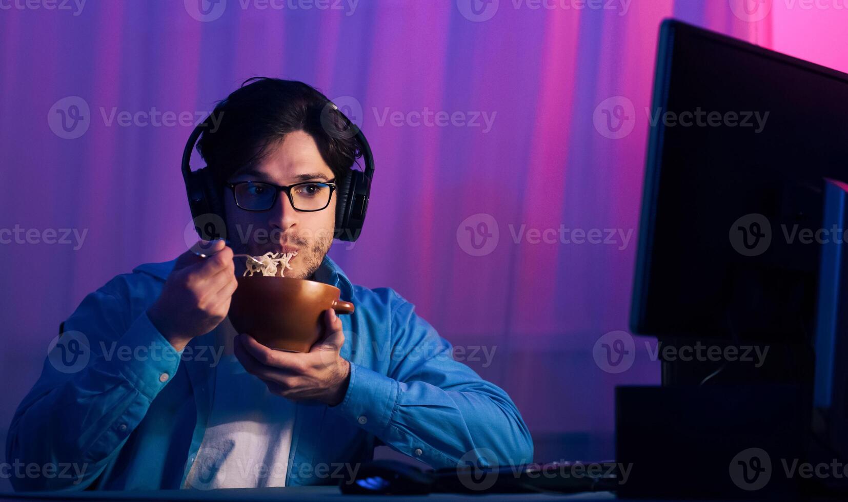 A young man sits in front of a computer screen, wearing headphones, while eating a bowl of noodles. He appears to be gaming at night, with colorful lights illuminating the background. photo