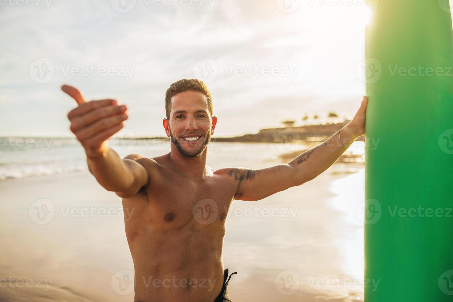 A young man stands on a sandy beach at sunset, holding a surfboard with his left hand and giving a thumbs up with his right. He is smiling and appears to be happy to be on the beach. photo