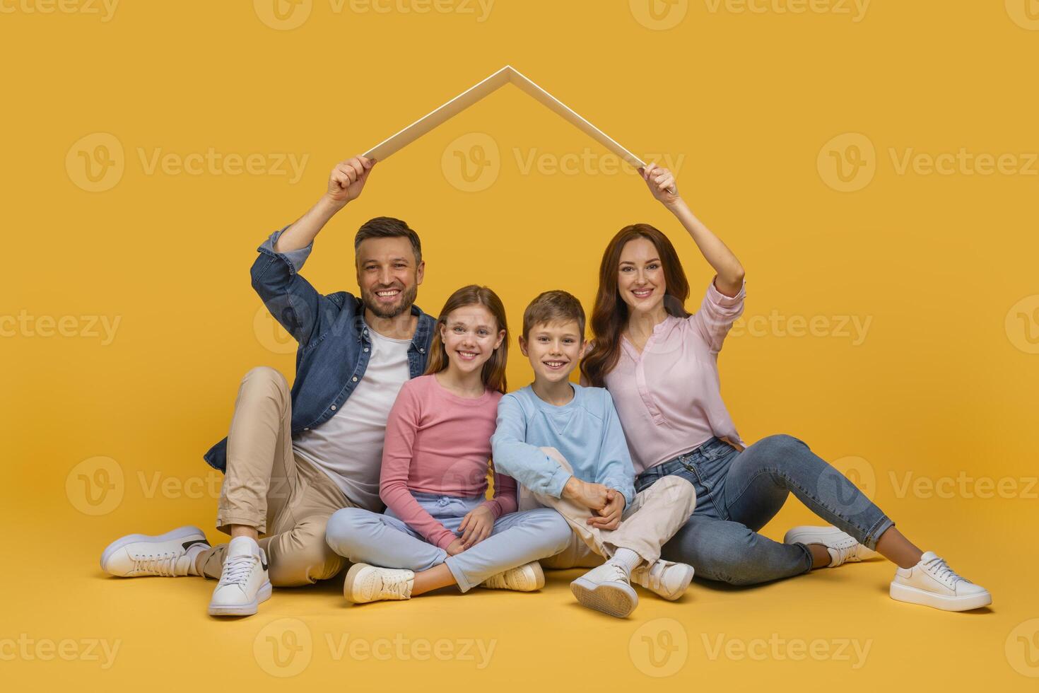 A happy family composed of a father, a mother, a daughter, and a son is sitting together on a yellow background, smiling as they hold a white picture frame above their heads photo