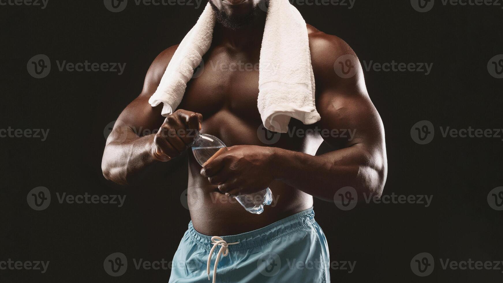 A muscular man wearing blue shorts and a white towel around his neck holds a water bottle after a strenuous workout. photo