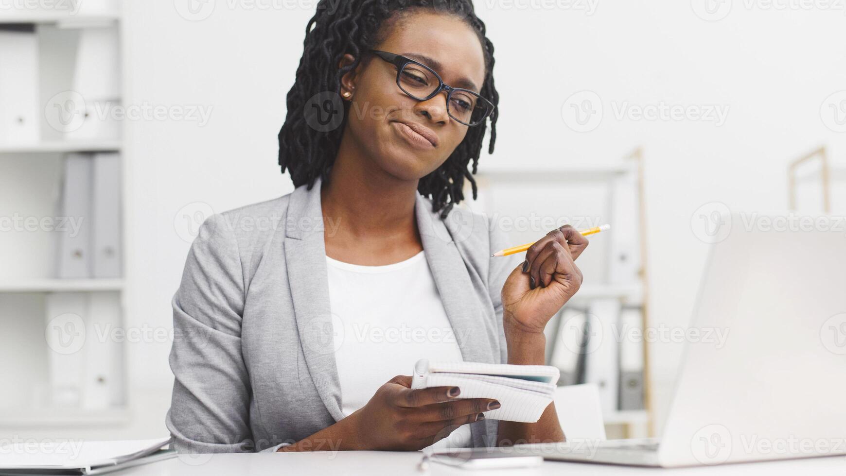 Thoughtful African American lady working on report, sitting at desk, writing in notepad, looking at laptop screen at modern office photo