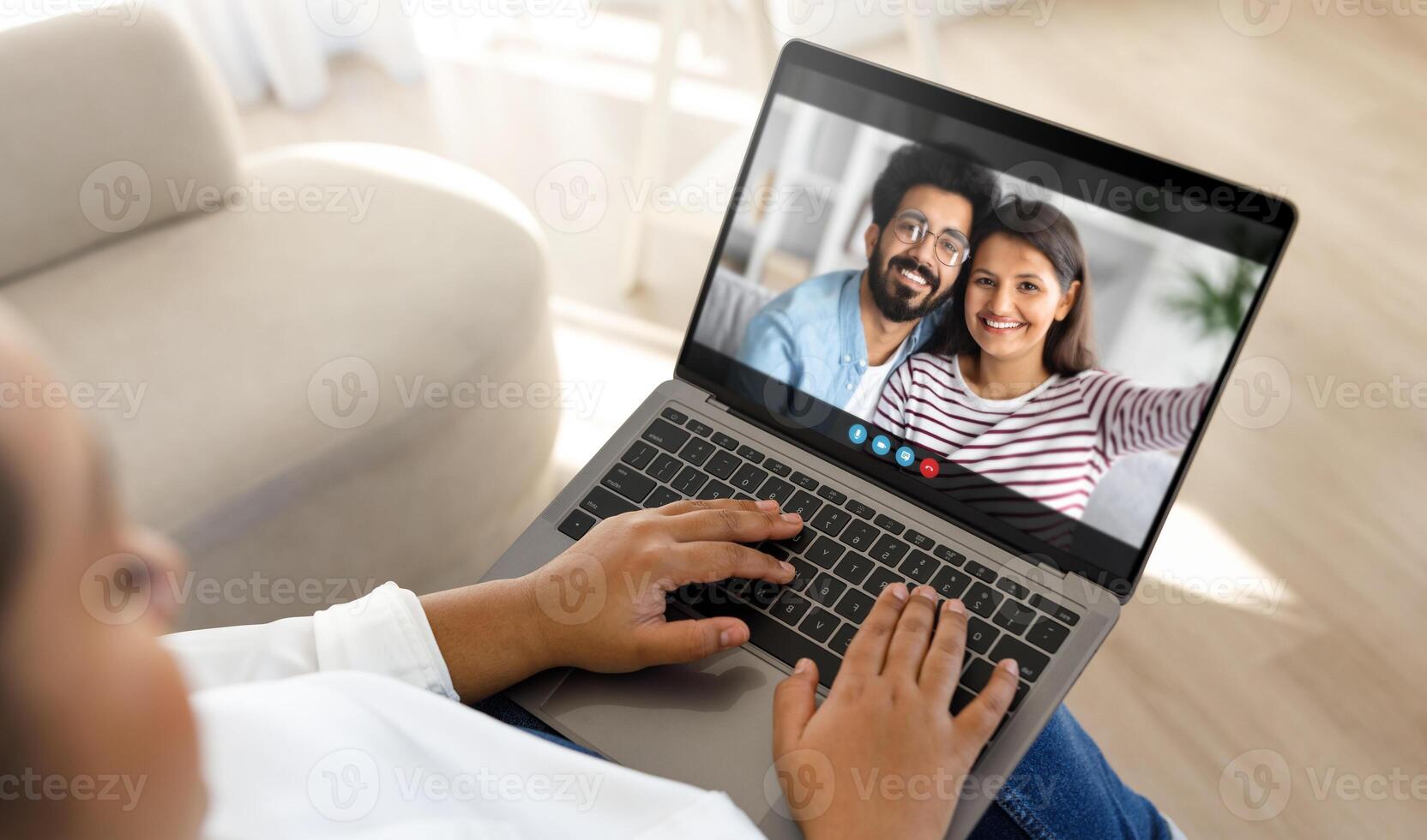Black lady person sits on a couch at home, using a laptop computer to chat with a couple. The person is typing on the laptop, while the couple smiles and waves at the camera. photo