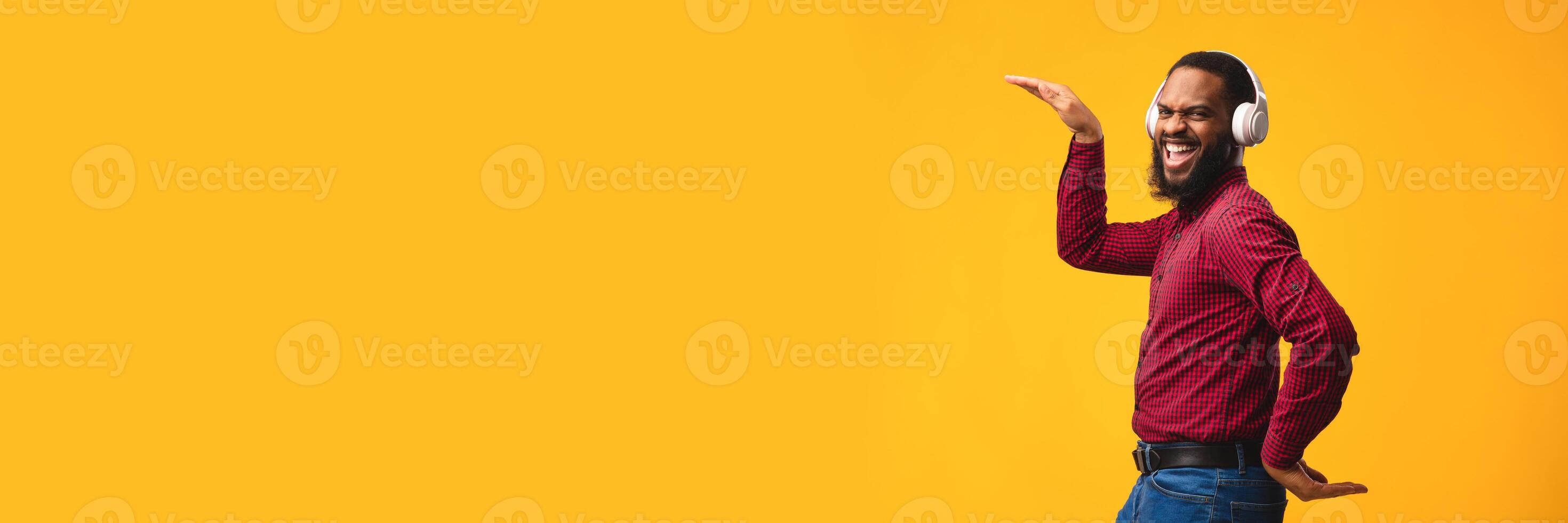 Enjoy The Sound. Happy black guy listening to music using headphones and dancing isolated over orange studio wall, panorama with copy space photo