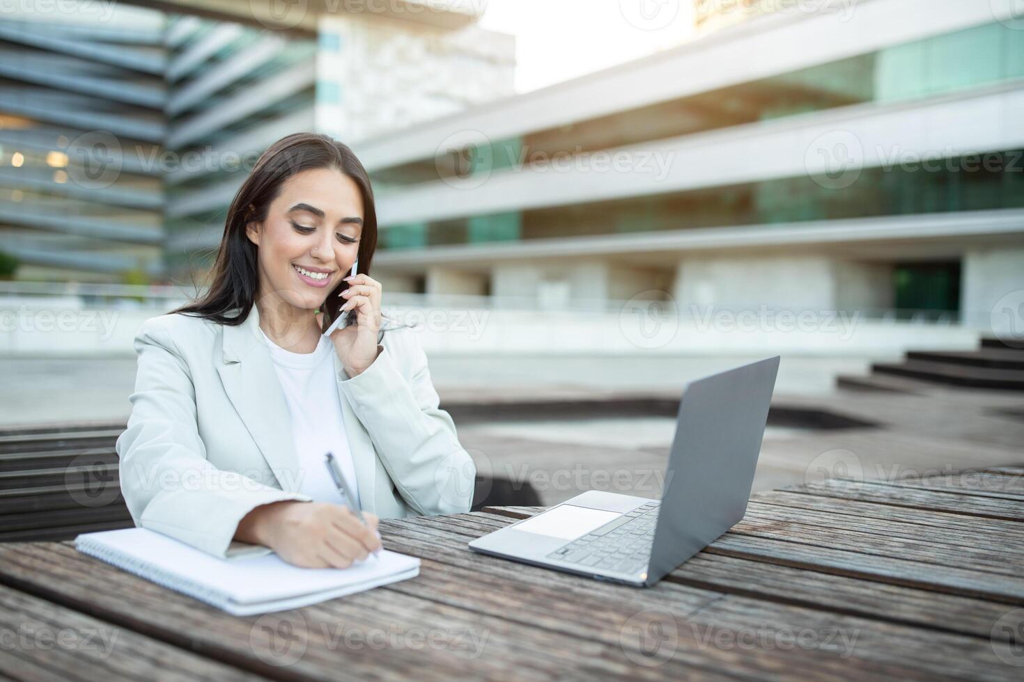 A young woman in a light blazer sits at a wooden table taking notes while speaking on her cellphone in an outdoor office setting. photo