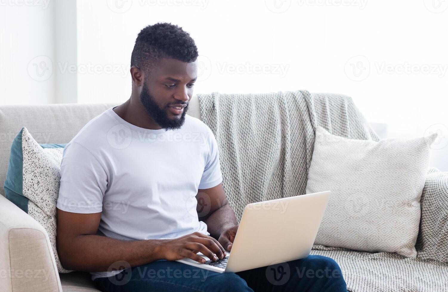 Black man in a white t-shirt sits on a couch in a living room, typing on a laptop computer. He has short dark hair, a beard, and is looking down at the computer screen. photo