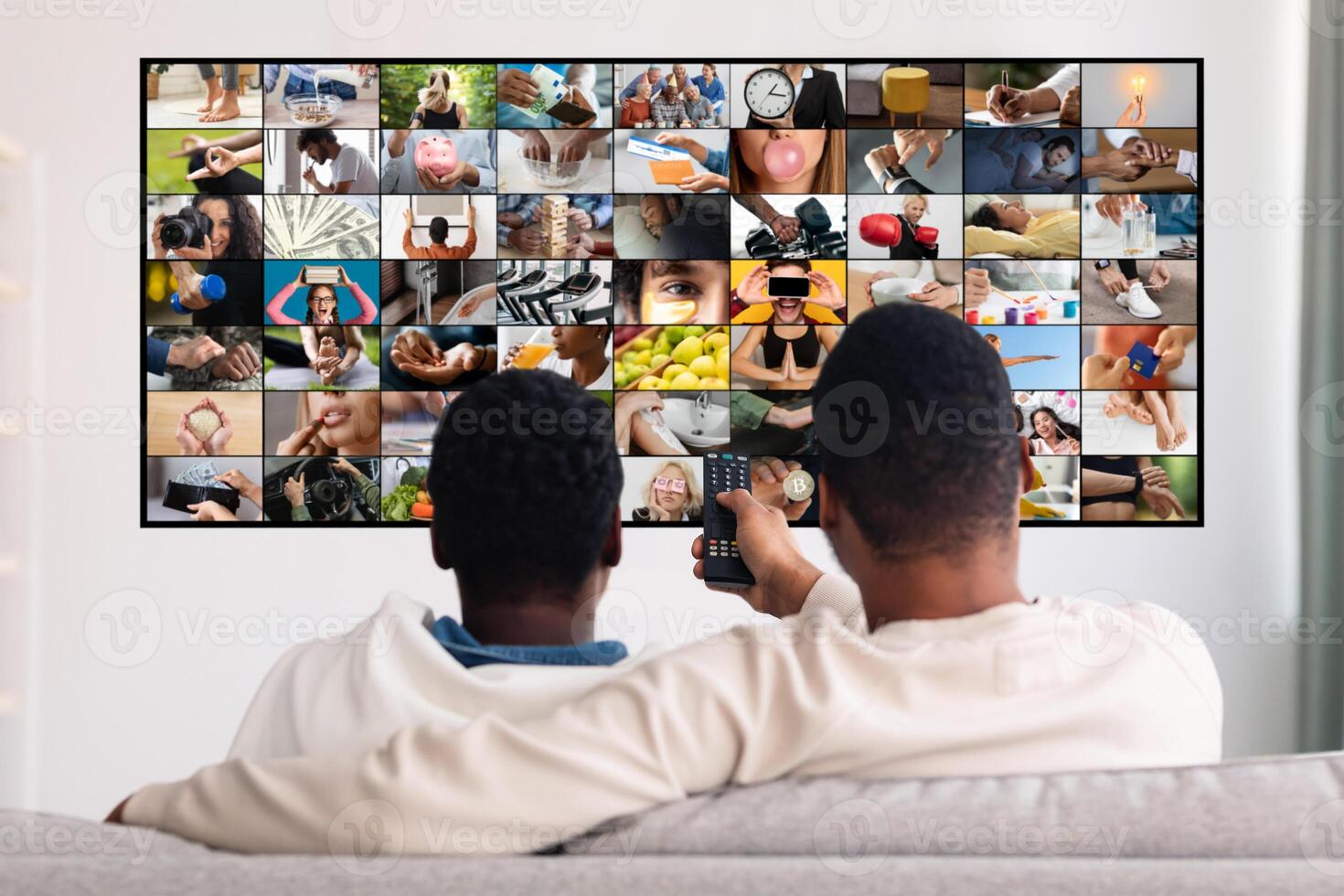Two men relax on a couch in their home watching television. They are both wearing casual clothing and appear to be enjoying their time together. Television screen displays a montage of various images photo