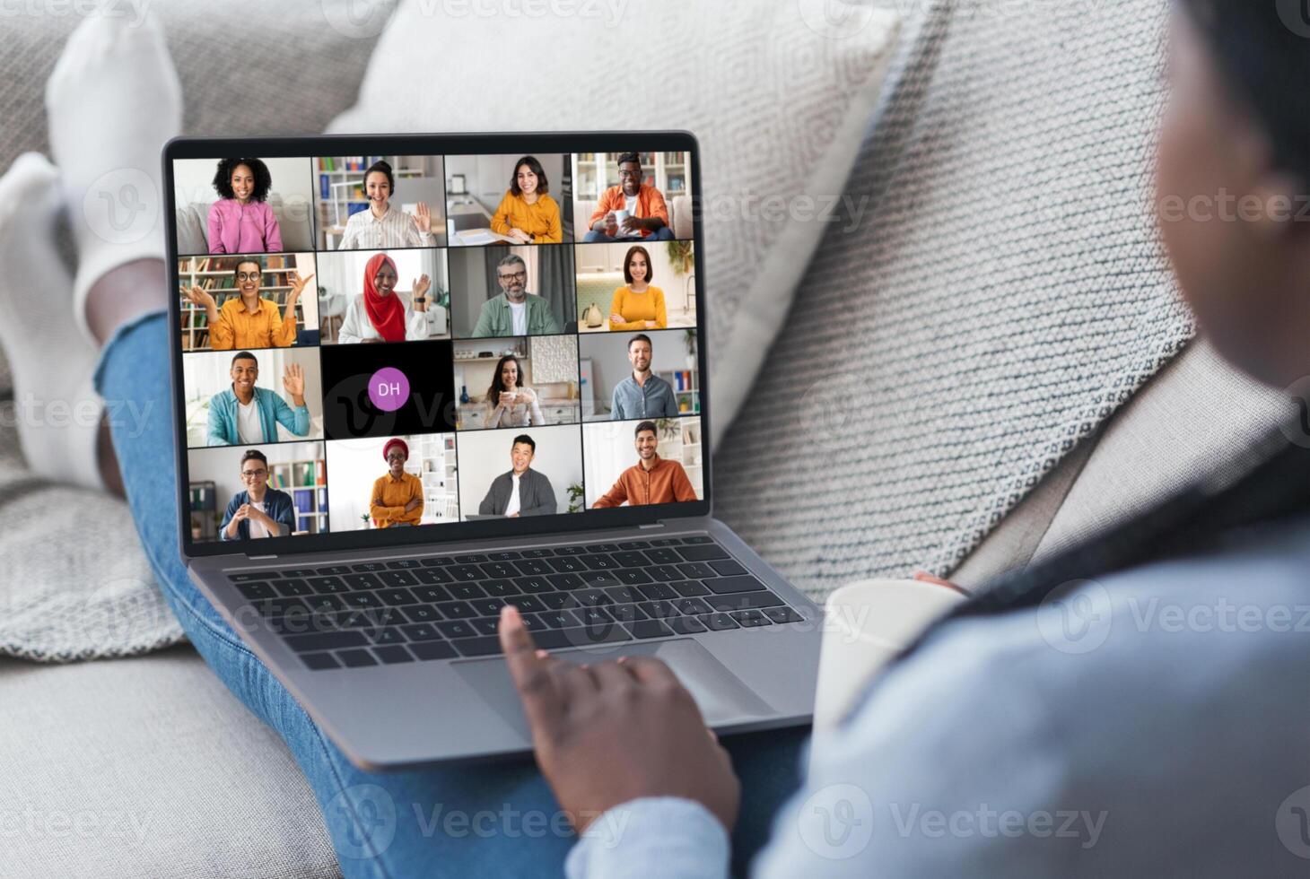 Black woman is sitting on a couch and participating in a conference with colleagues. She is looking at the laptop screen, which shows a grid of feeds of the other participants. photo