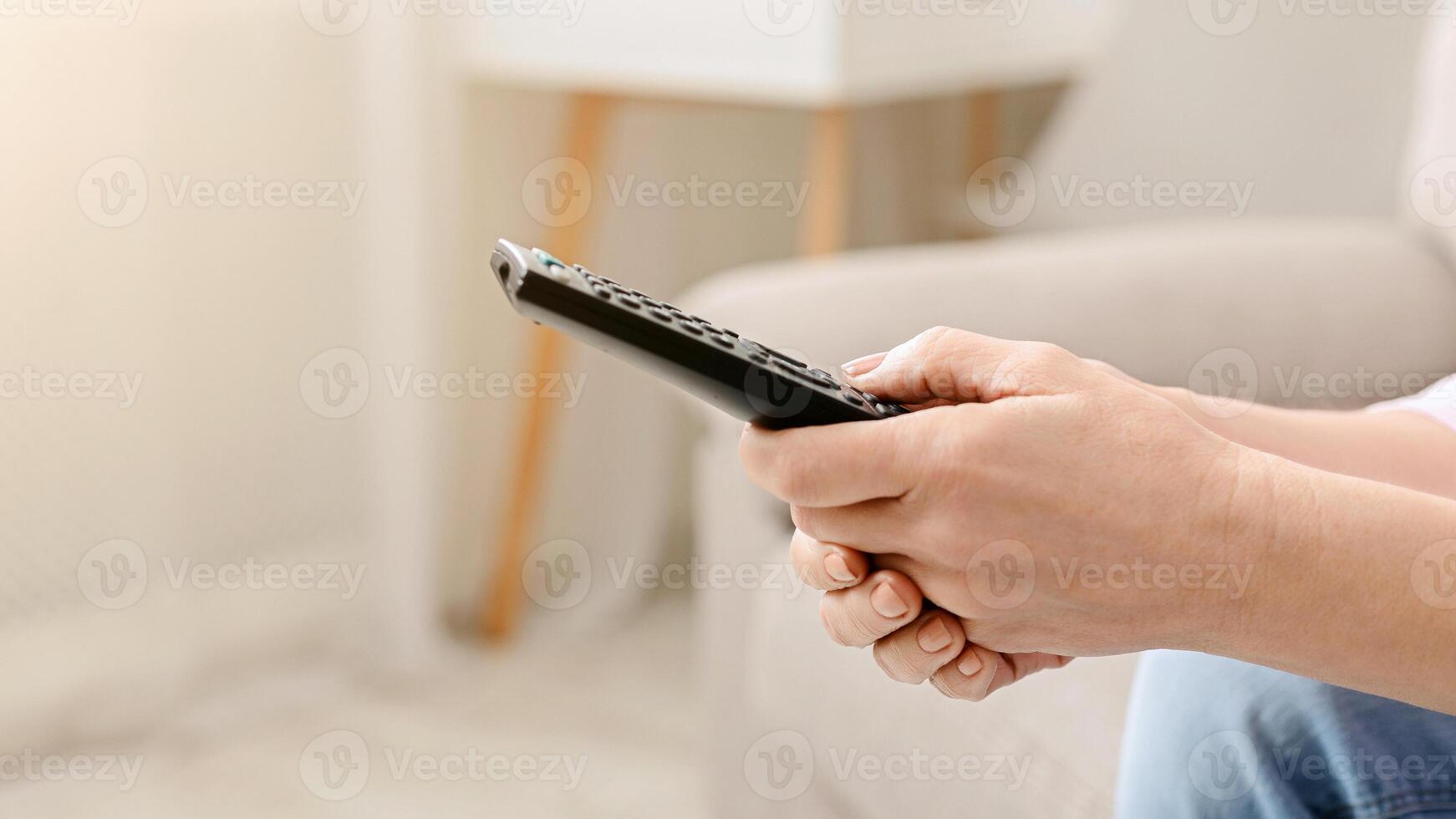 Cropped of woman is seated on a couch, holding a remote control in hand, appear to be watching television or adjusting settings on a device photo