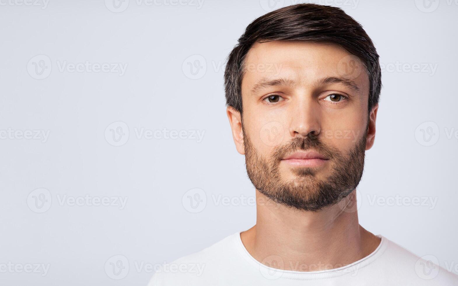 A man with a beard looks directly at the camera against a white background. He is wearing a white t-shirt. photo