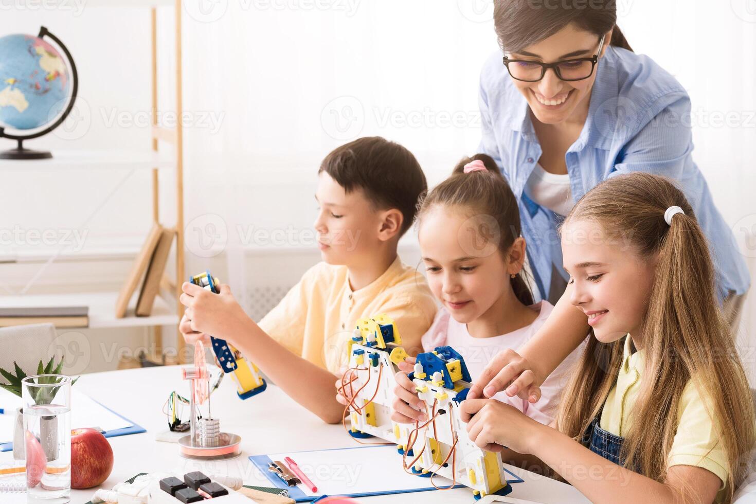 A group of elementary school students are working on a robotics project in a classroom setting. They are building robots with the help of their teacher, who is smiling and guiding them. photo