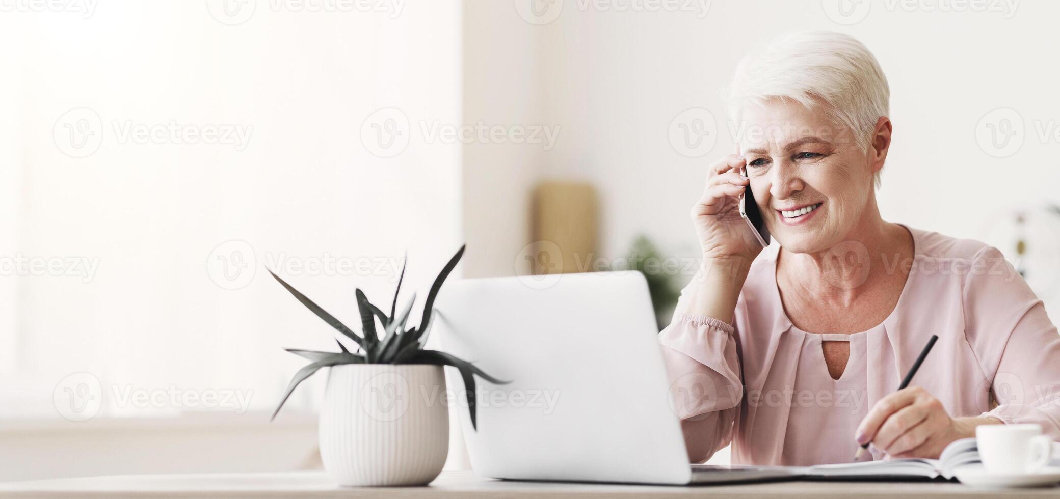 Smiling elderly woman talking to business partners by phone, looking at laptop, making notes, copy space photo