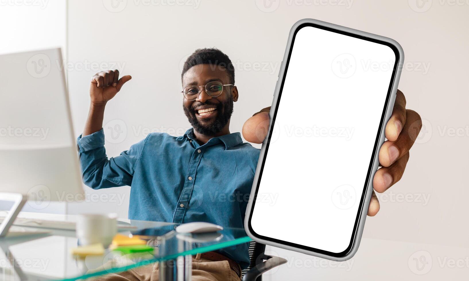 African American man sitting at workdesk with pc, holding a cell phone in his hand, showing blank white screen and gesturing photo