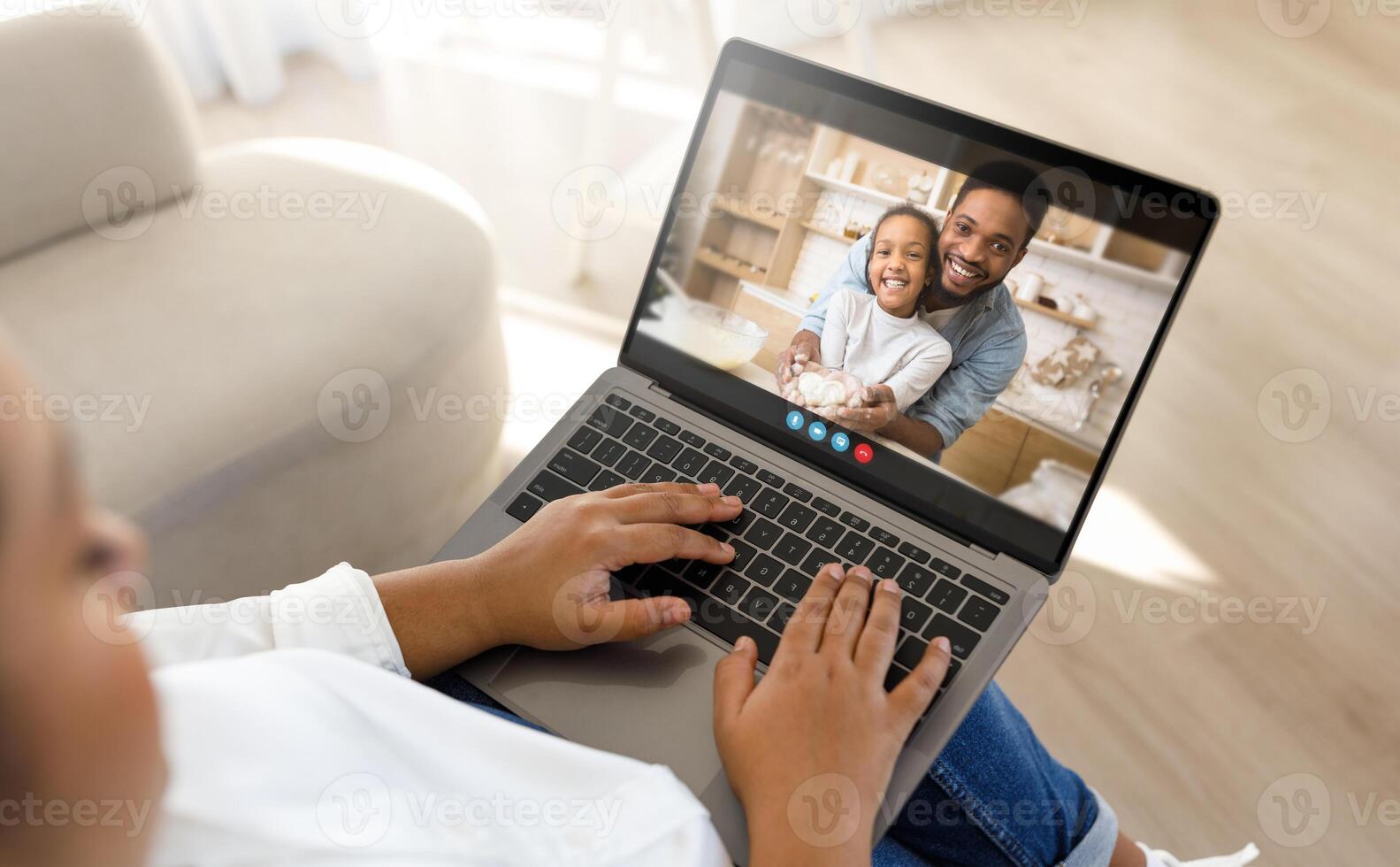 Black woman mother sits on a couch and uses a laptop to chat with their family. The person on the laptop screen is a man and a young girl, both smiling and looking happy. photo