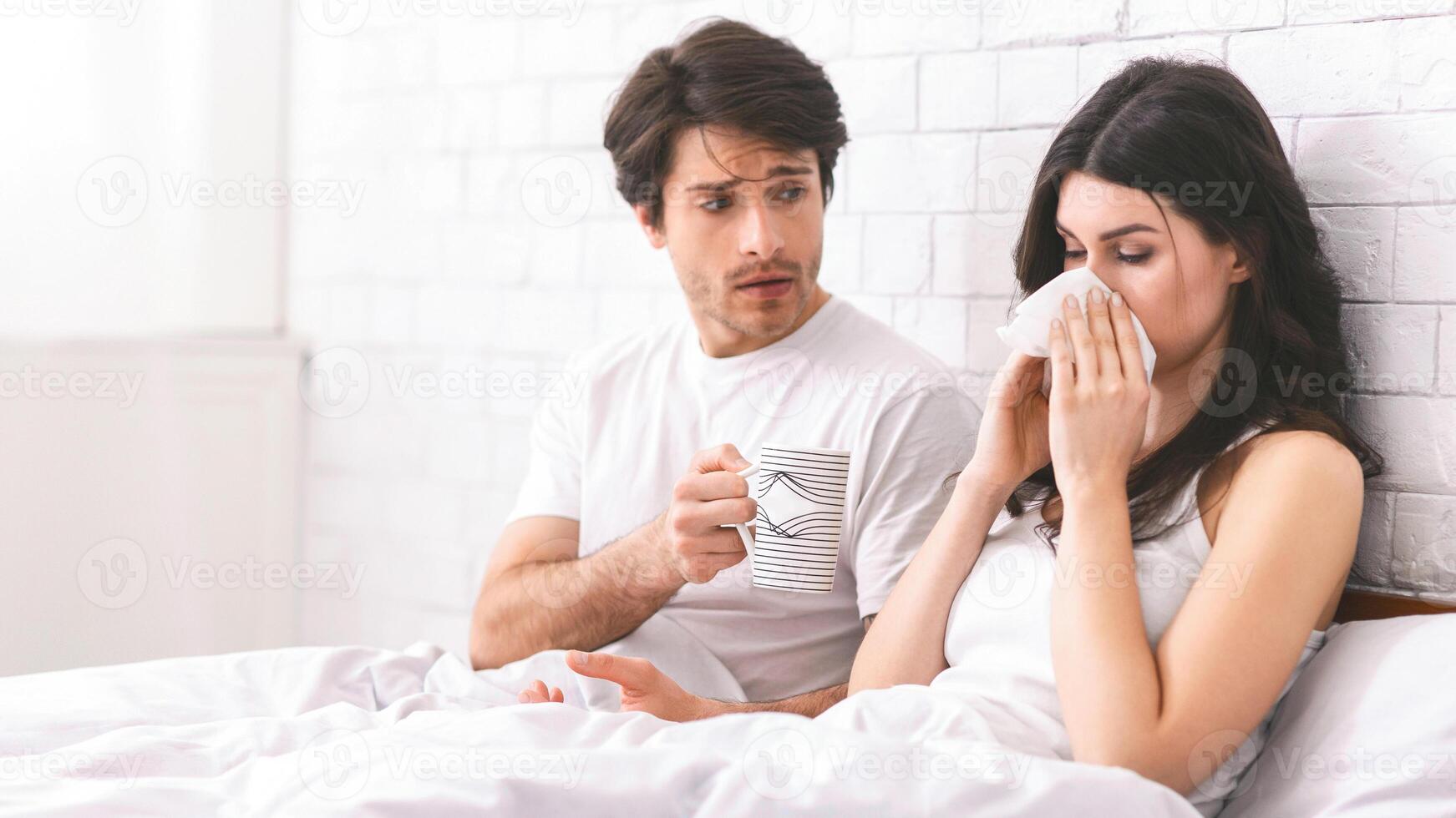 A man with a worried expression holds a mug of tea while a sick woman blows her nose with a tissue in bed. The couple are in a bedroom with a white brick wall. photo