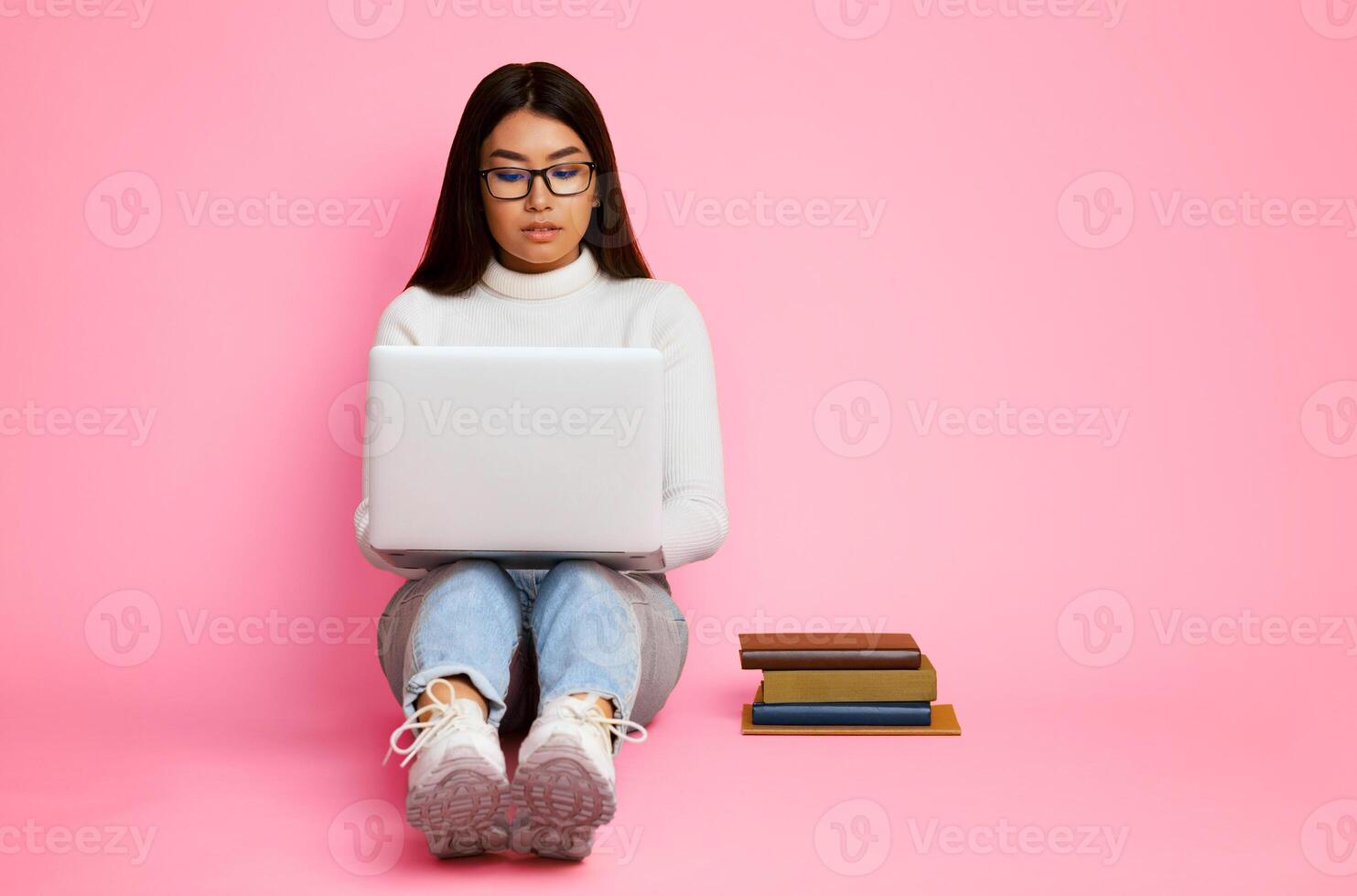 Asian young woman sitting on a pink background while using a laptop. She is looking at the laptop screen and typing. There are three books stacked next to her, copy space photo