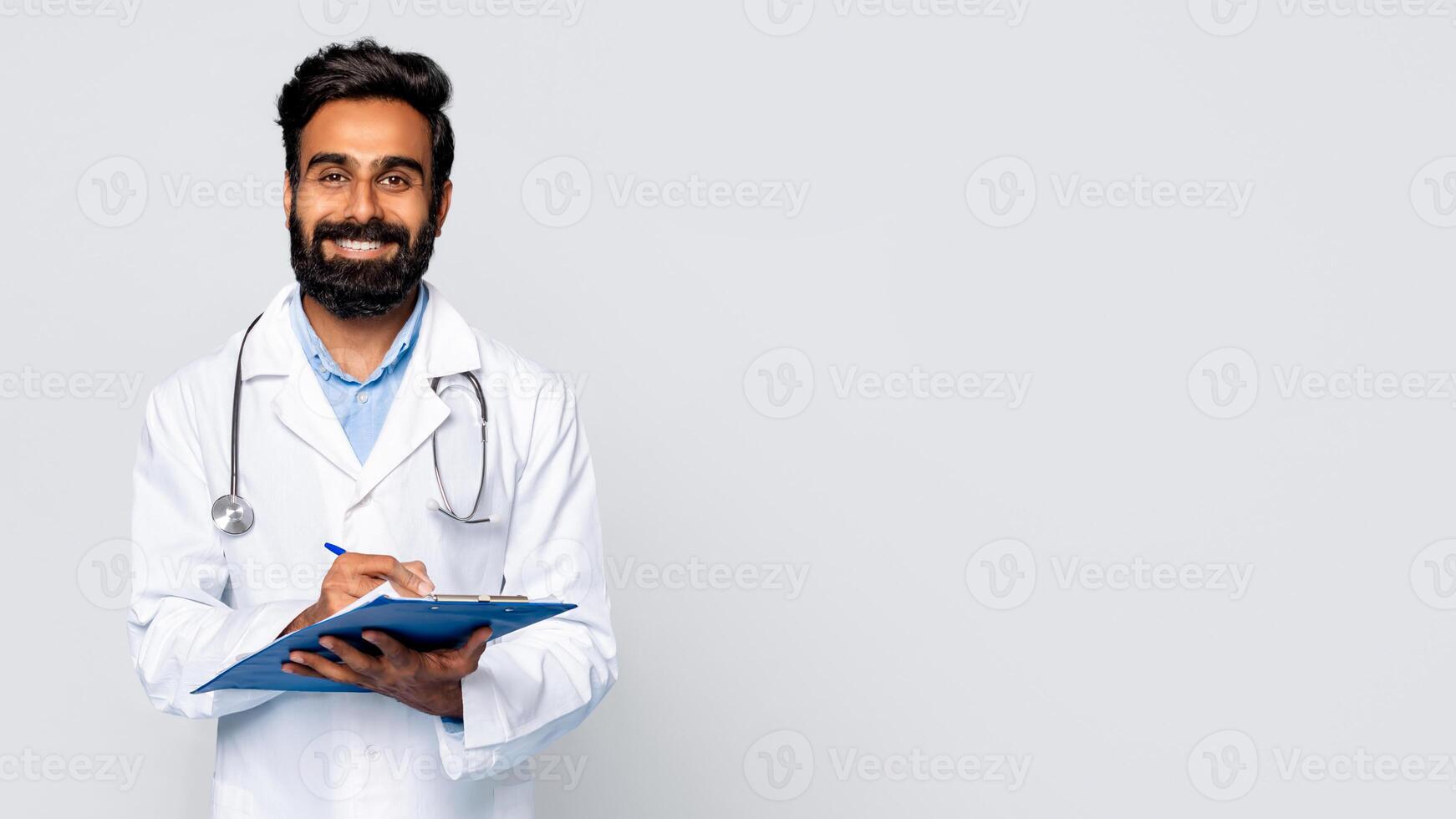 Eastern man wearing a white lab coat and a stethoscope smiles while writing on a blue clipboard with a pen. The background is a plain, light gray, copy space photo