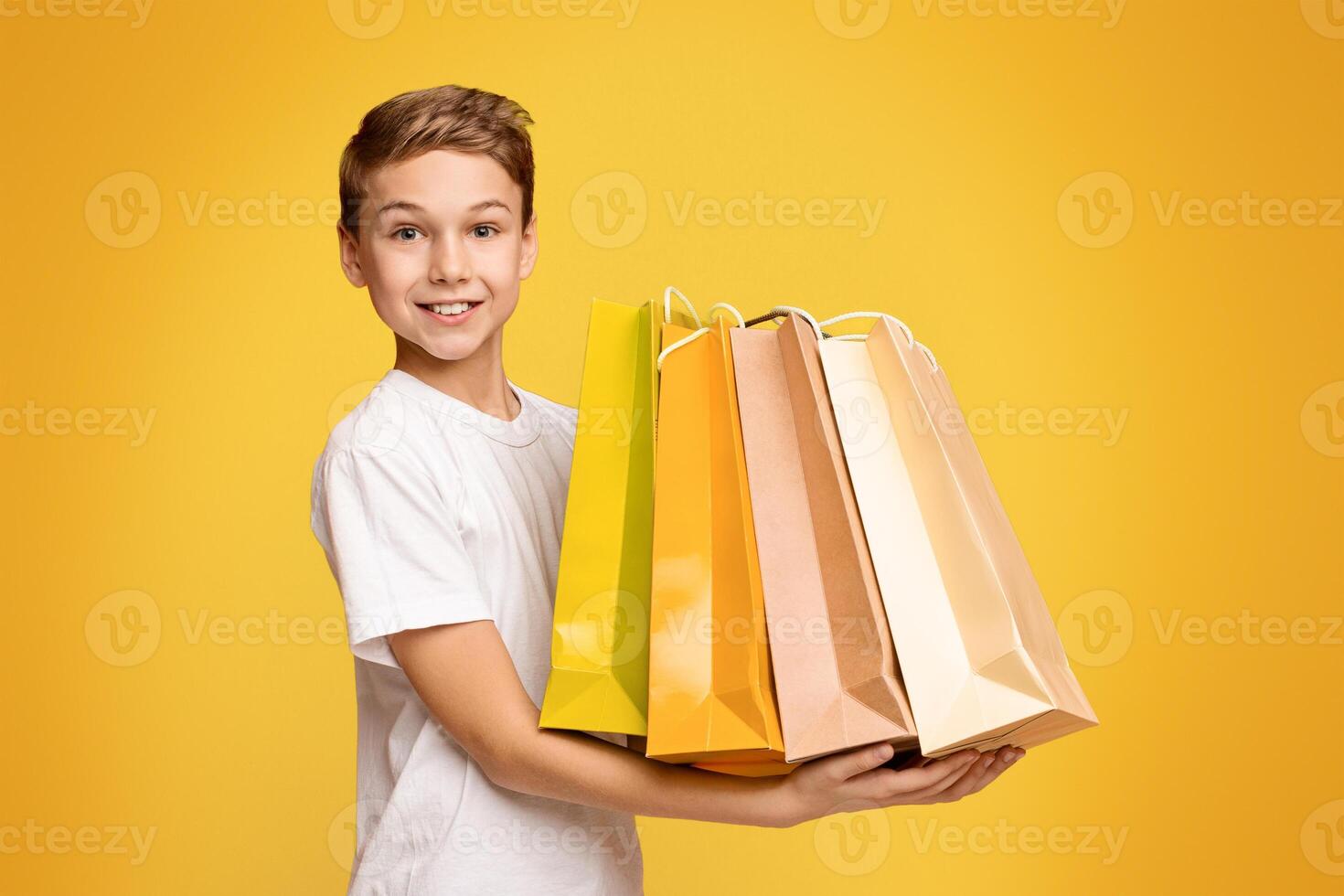 Little shopper. Happy teen boy holding lots of colorful paper shopping bags in hands, orange studio background photo