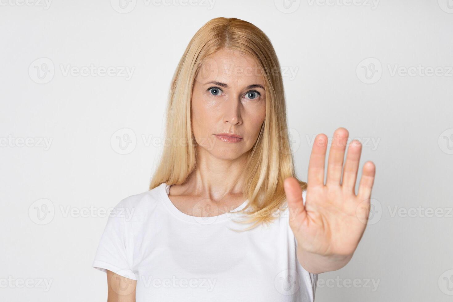 A blonde woman wearing a white t-shirt is looking directly at the camera with a serious expression. She is holding her right hand up, palm facing forward, in a clear stop gesture photo