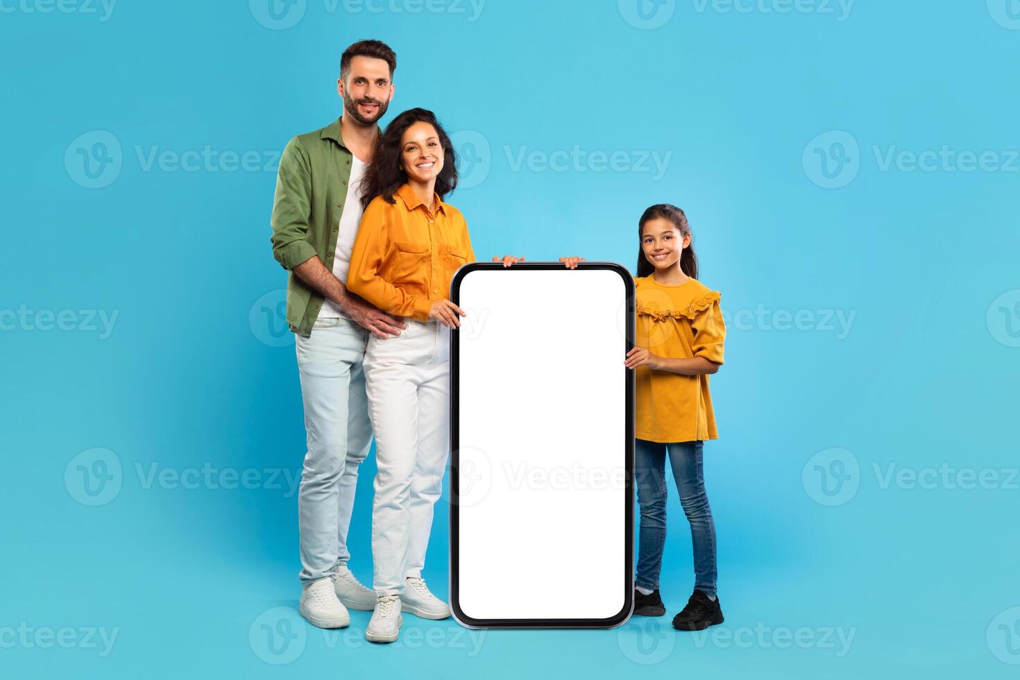 A smiling family, consisting of a father, mother, and daughter, stand together in front of a blue background. The parents are holding a large smartphone with a blank screen photo
