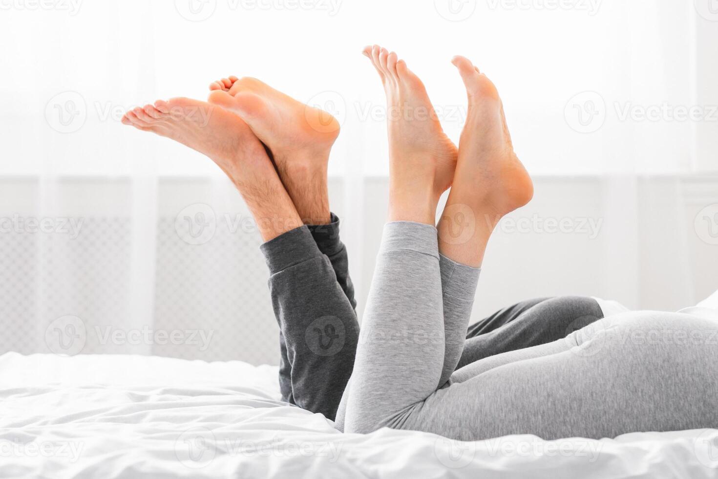 A photo of a couples feet raised in the air as they lie in bed together. The soft white bedding and natural light streaming in through the window create a peaceful and intimate atmosphere.