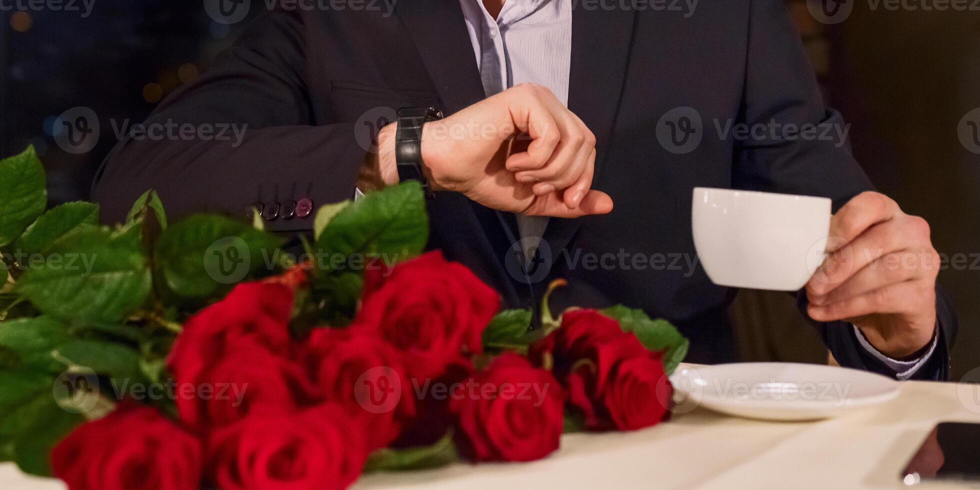 Man with flowers waiting girlfriend in restaurant nervous, drinking coffee, checking time. She is late concept photo