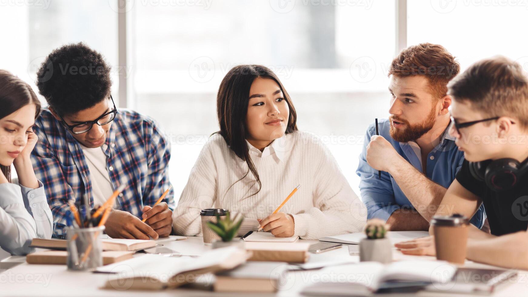 A diverse group of students sitting together at a table, engaged in conversation and possibly doing homework. Some are gesturing while speaking, while others are listening attentively photo