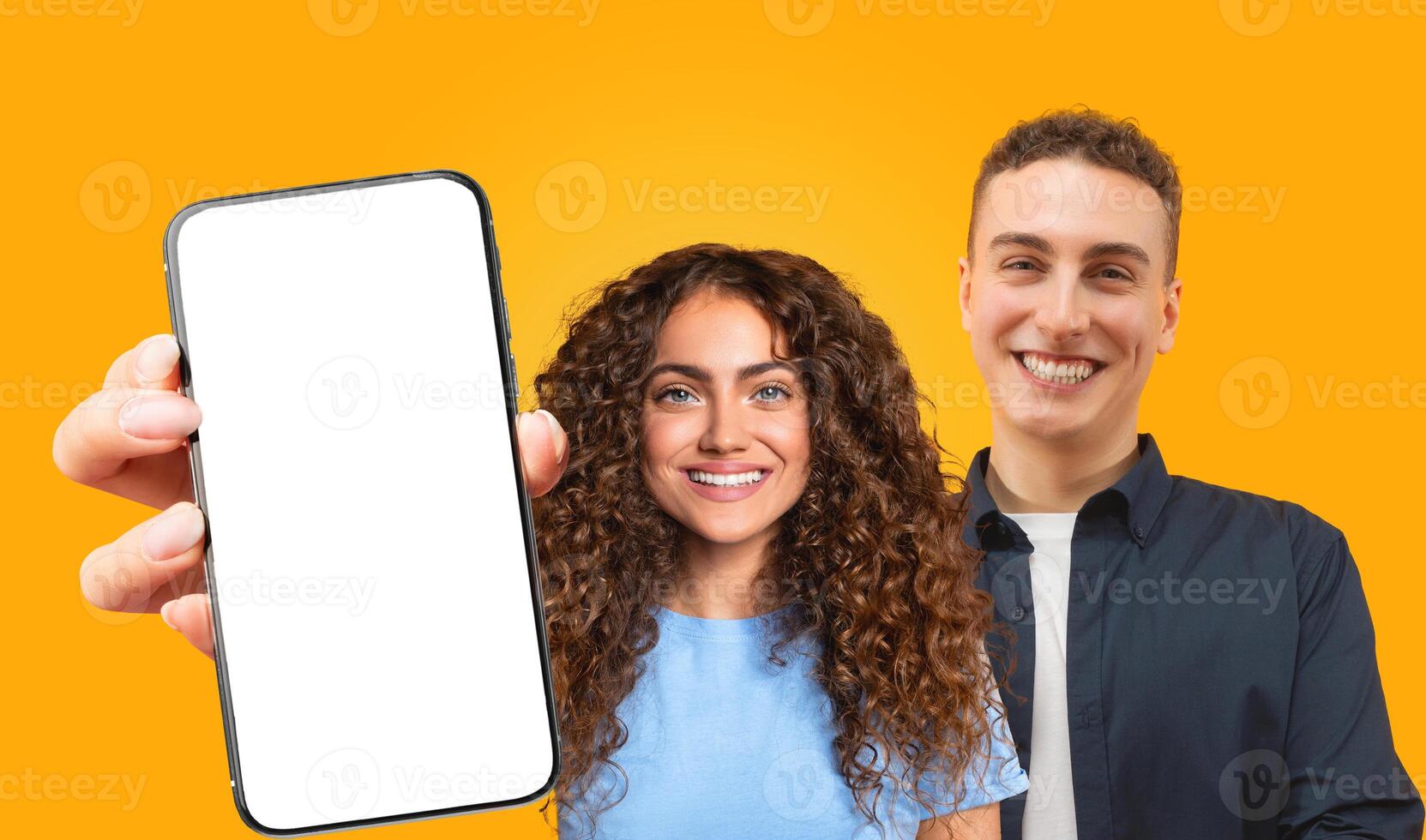 A young woman with long curly brown hair holds a smartphone with a blank white screen in front of her, smiling at the camera. She is standing next to a young man with short brown hair photo