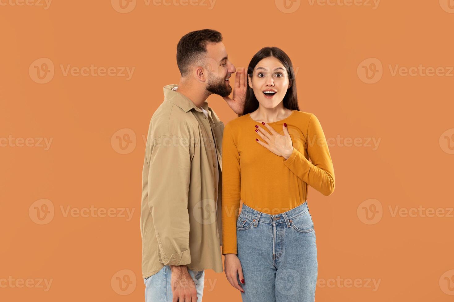 A man leans in to whisper a secret to a surprised and delighted woman. They stand against a vibrant orange background, highlighting their casual clothing and excited expressions photo