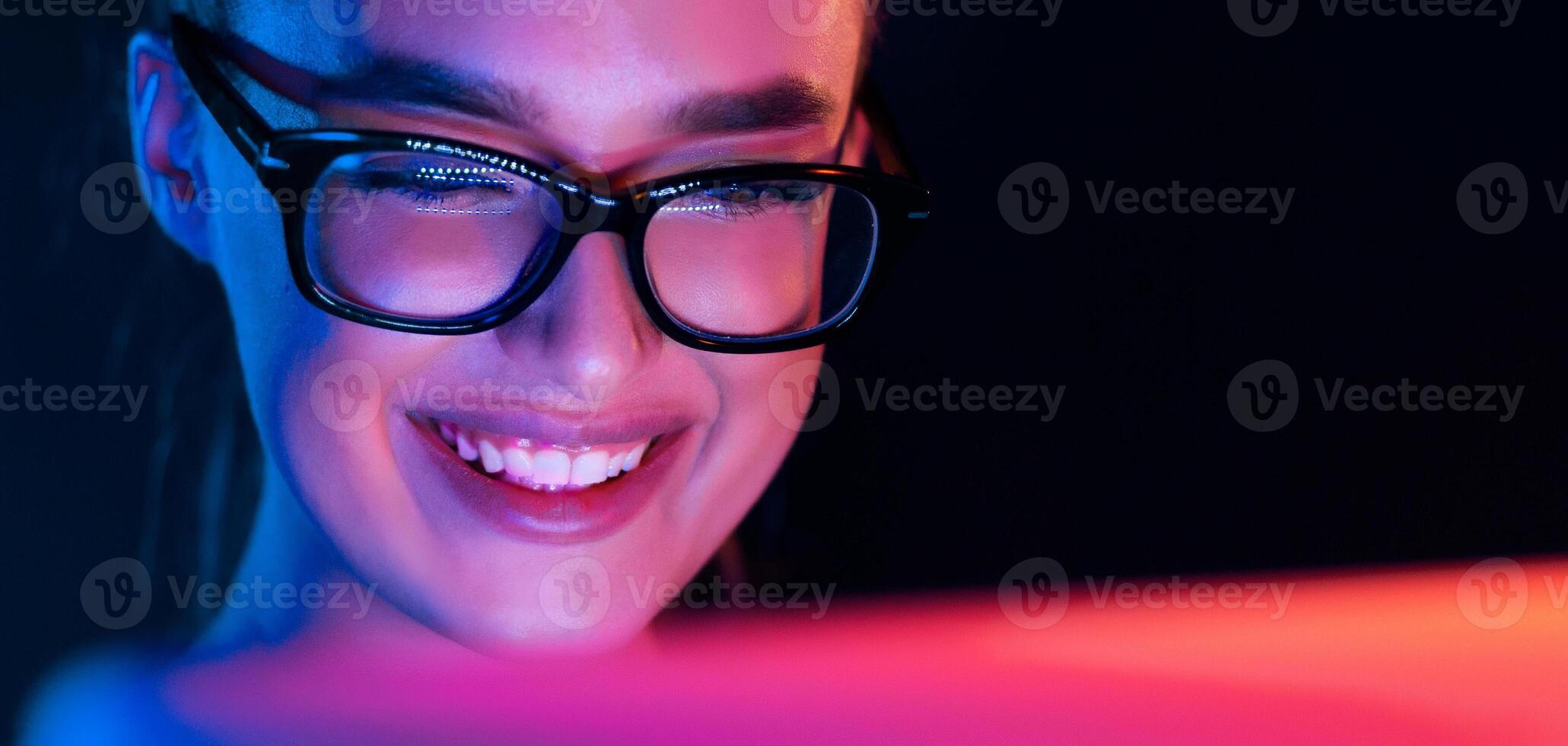 A woman wearing glasses is sitting in front of a computer screen, smiling brightly as she looks at the display. Her expression conveys happiness and engagement with the content on the screen. photo