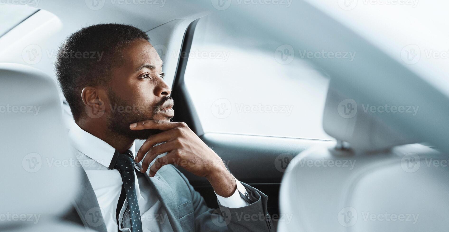African American businessman in a grey suit, white shirt, and patterned tie sits in the back seat of a car. He is looking out the window and appears to be deep in thought, copy space photo