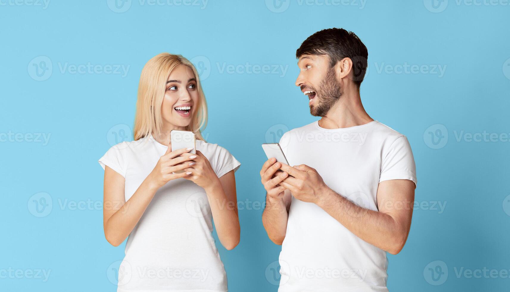 A young woman and man are standing in front of a blue background, both holding smartphones. The woman is looking at the man's phone with a surprised expression photo
