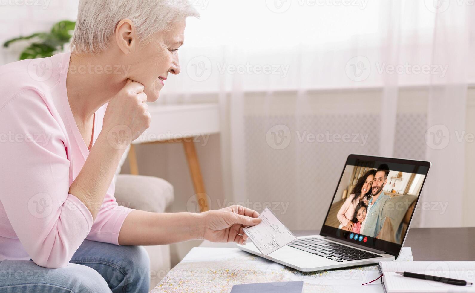 A senior woman is sitting at a table with a laptop in front of her. She is smiling and holding up a check for her family on the call. The family is smiling back at her. photo
