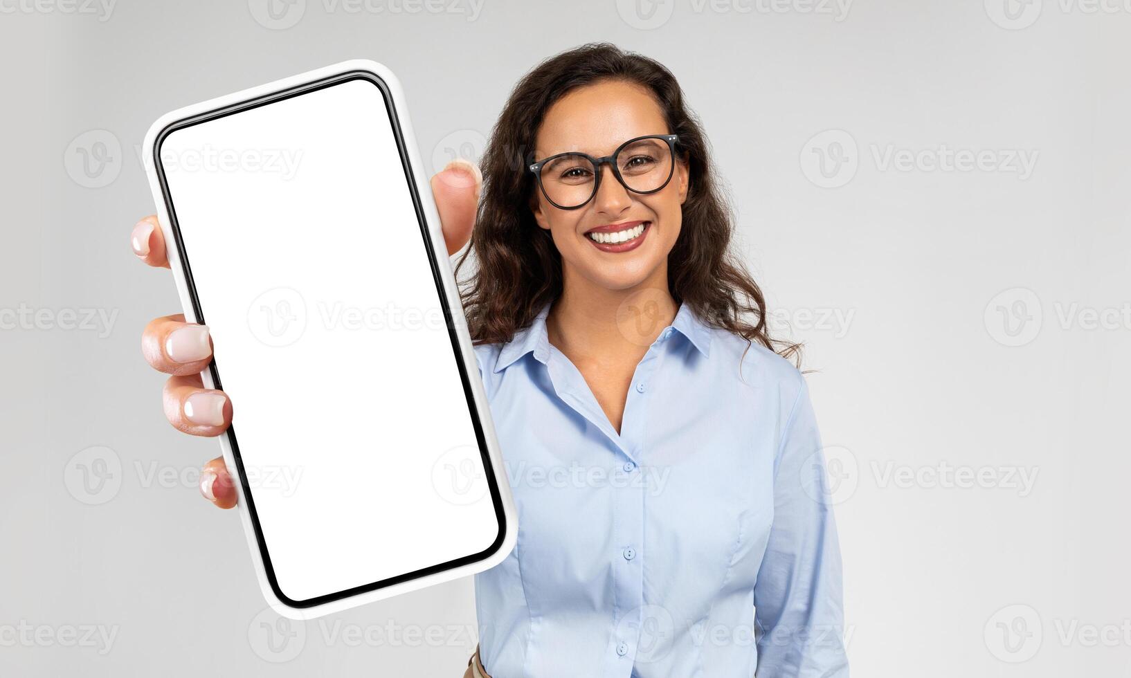 Businesswoman is seen holding a modern cell phone in her hand. The phones screen is blank, showing no content. She appears to be focused on the device, possibly waiting for a message or browsing. photo