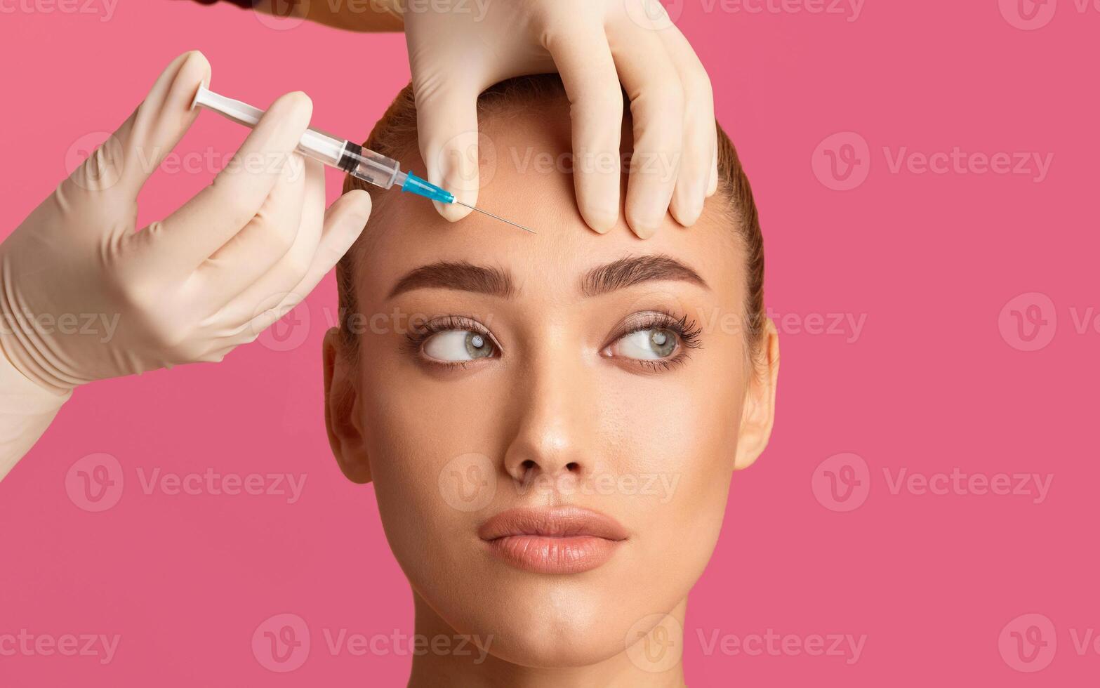 A woman receives a forehead injection treatment from a medical professional wearing gloves. The image is taken against a pink background. The womans expression is relaxed and she is looking upwards. photo