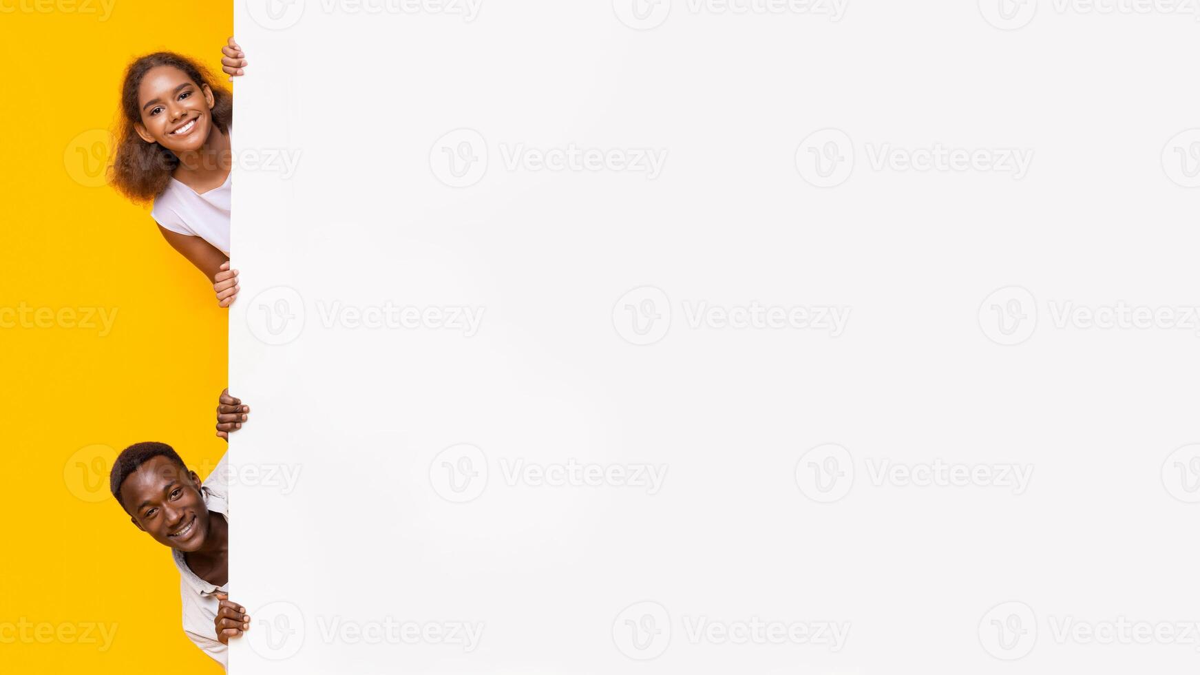 African boy and girl looking from behind blank advertising board, yellow background photo