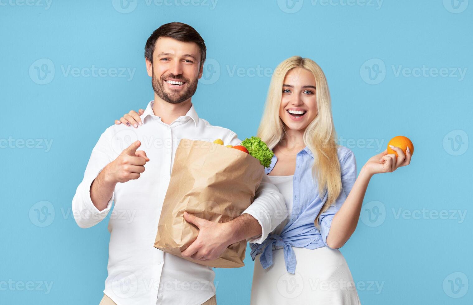 A man and a woman stand together against a blue background. The man is holding a brown paper bag filled with groceries and pointing to the side with his right hand photo