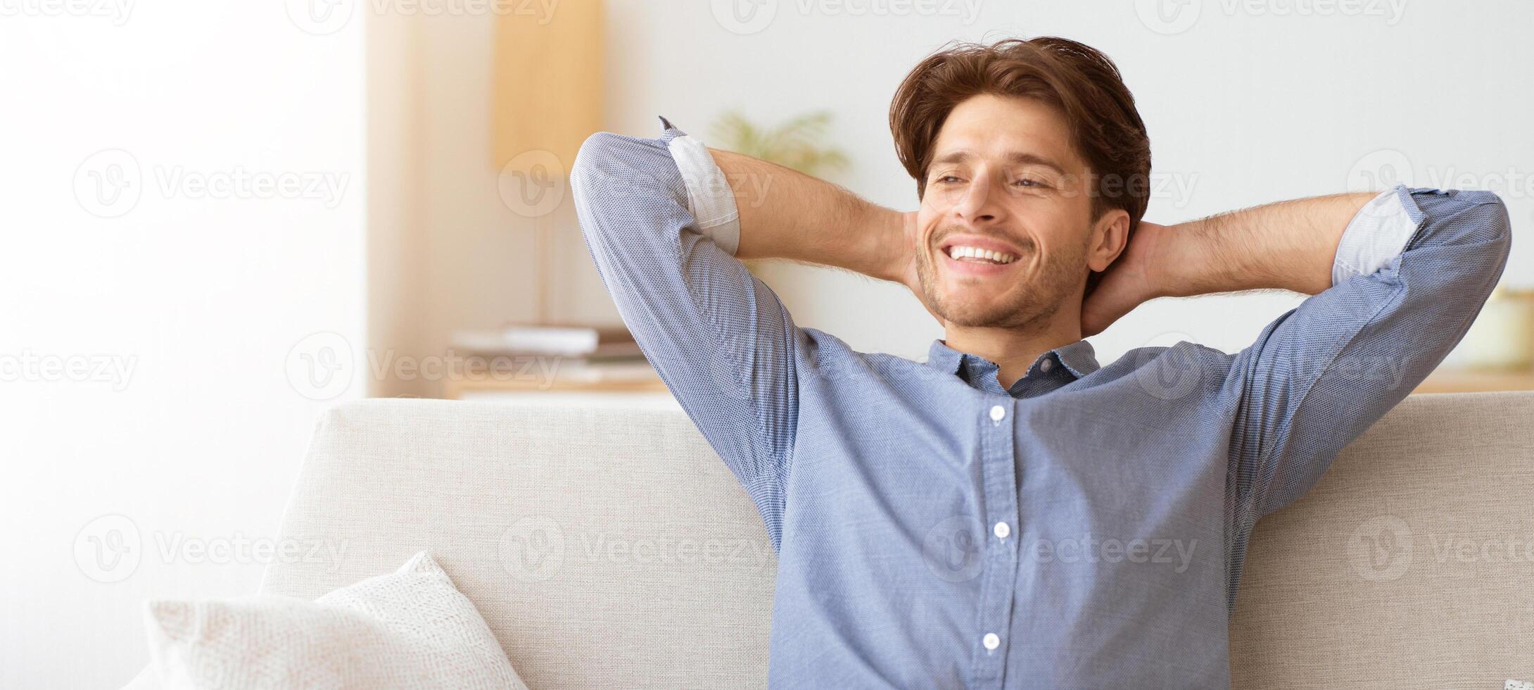 A young man sits on a couch with his arms behind his head, smiling and looking relaxed. photo