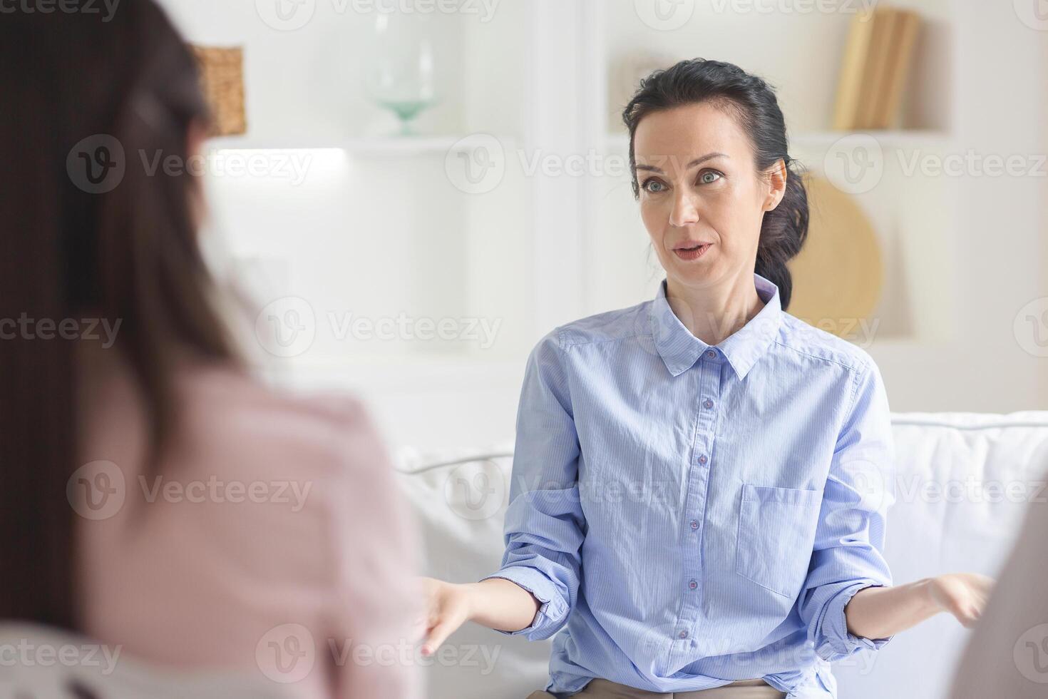 A woman in a light blue shirt is engaging in a counseling session during daytime in a cozy, well-lit office with white walls and minimal decor. She appears attentive and expressive photo