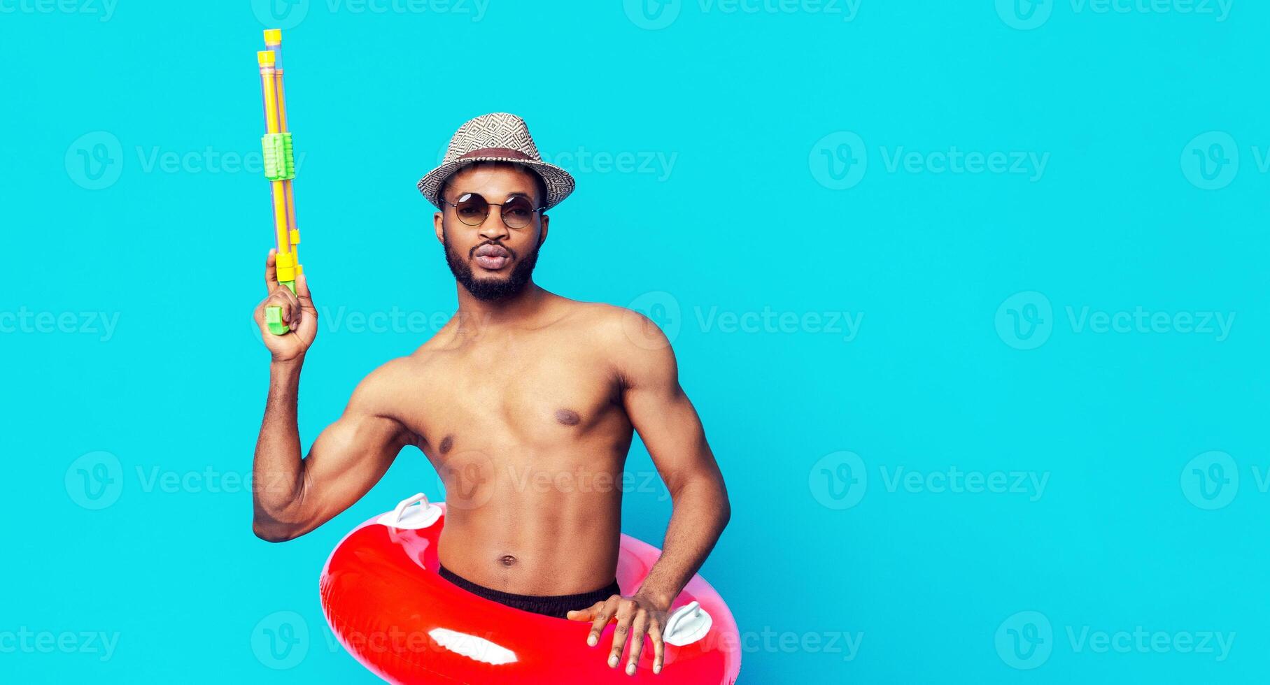 African American young man wearing a hat and sunglasses holds a water gun while standing in front of a blue background. He is wearing a life preserver around his waist. photo