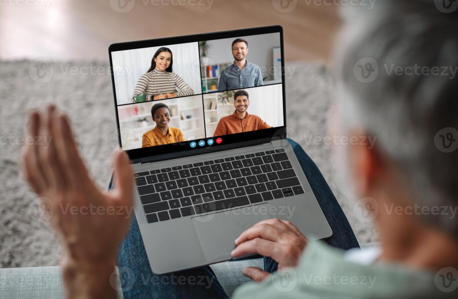 Senior man sits on a couch, his back to the camera, actively participating in a conference call on his laptop. Screen displays four participants, all smiling and engaging in the virtual meeting photo
