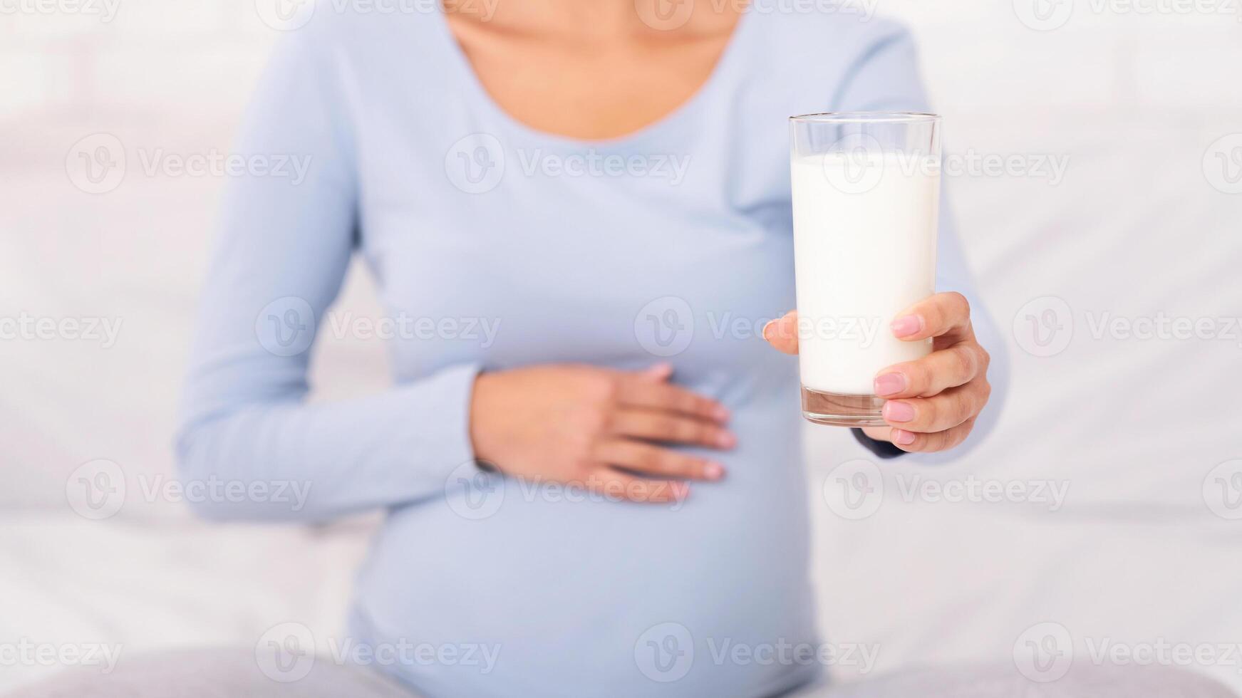 Dairy In Pregnancy Diet. Unrecognizable Pregnant Woman Offering Glass Of Milk To Camera Sitting On Bed In Bedroom. Cropped photo