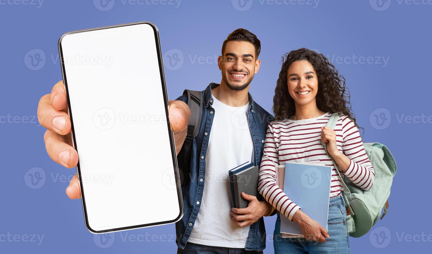 Two arabic students are standing in front of a blue background. They are both smiling and holding books and backpacks. The man is holding a smartphone with a blank screen in front of him. photo