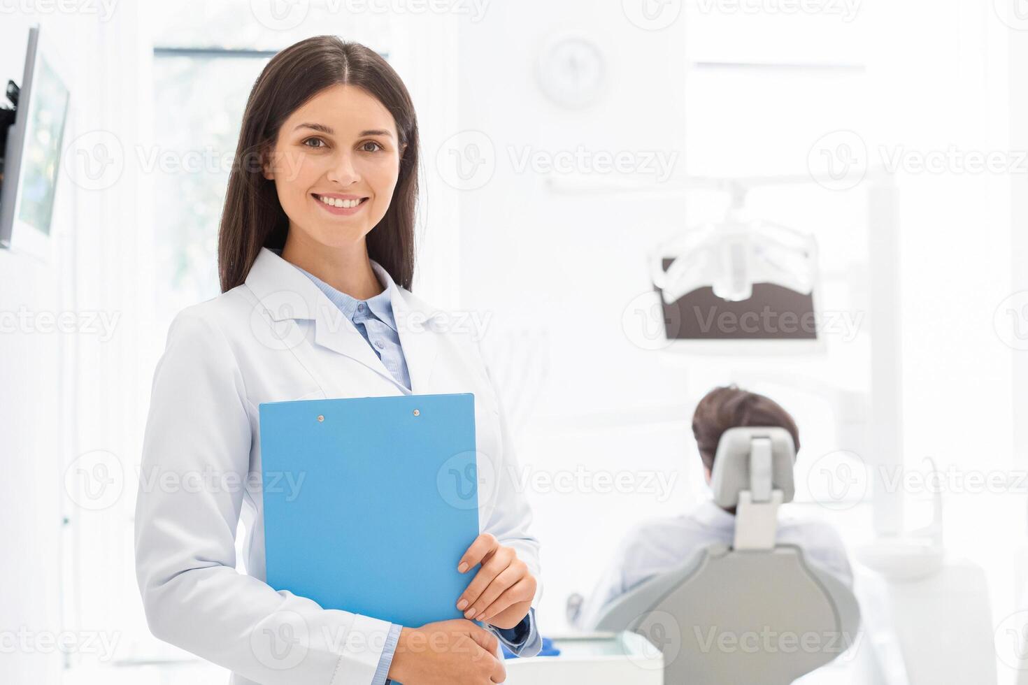 Attractive young female dentist smiling at camera, holding folder over dental office background, copy space photo