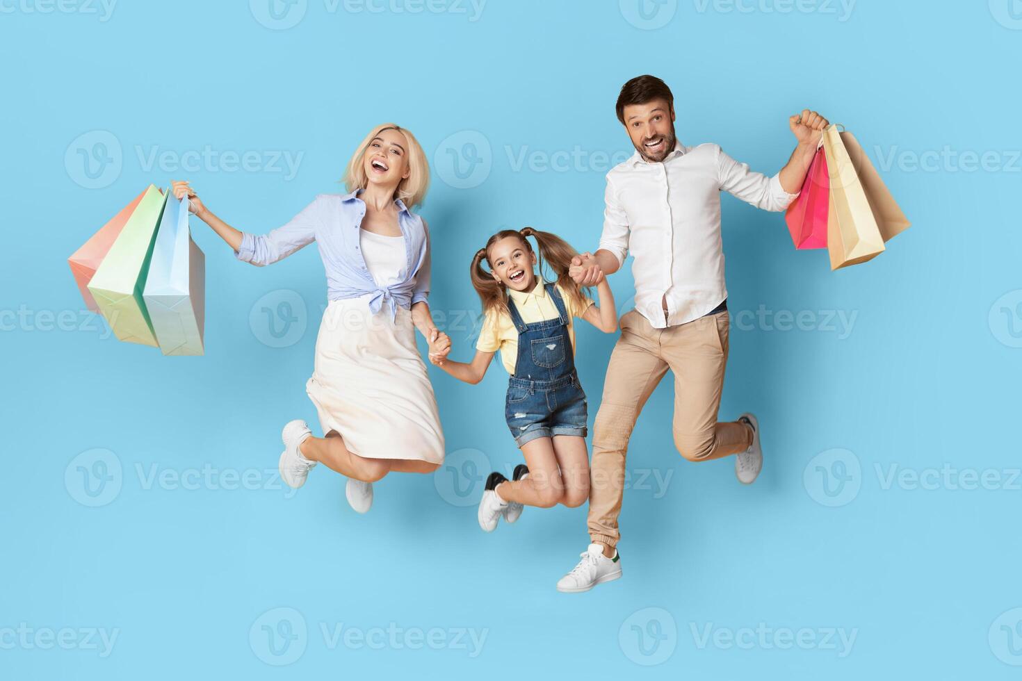 A happy family of three, a mother, father, and young daughter, all wearing casual clothes and holding shopping bags, jump in the air in front of a blue background. photo
