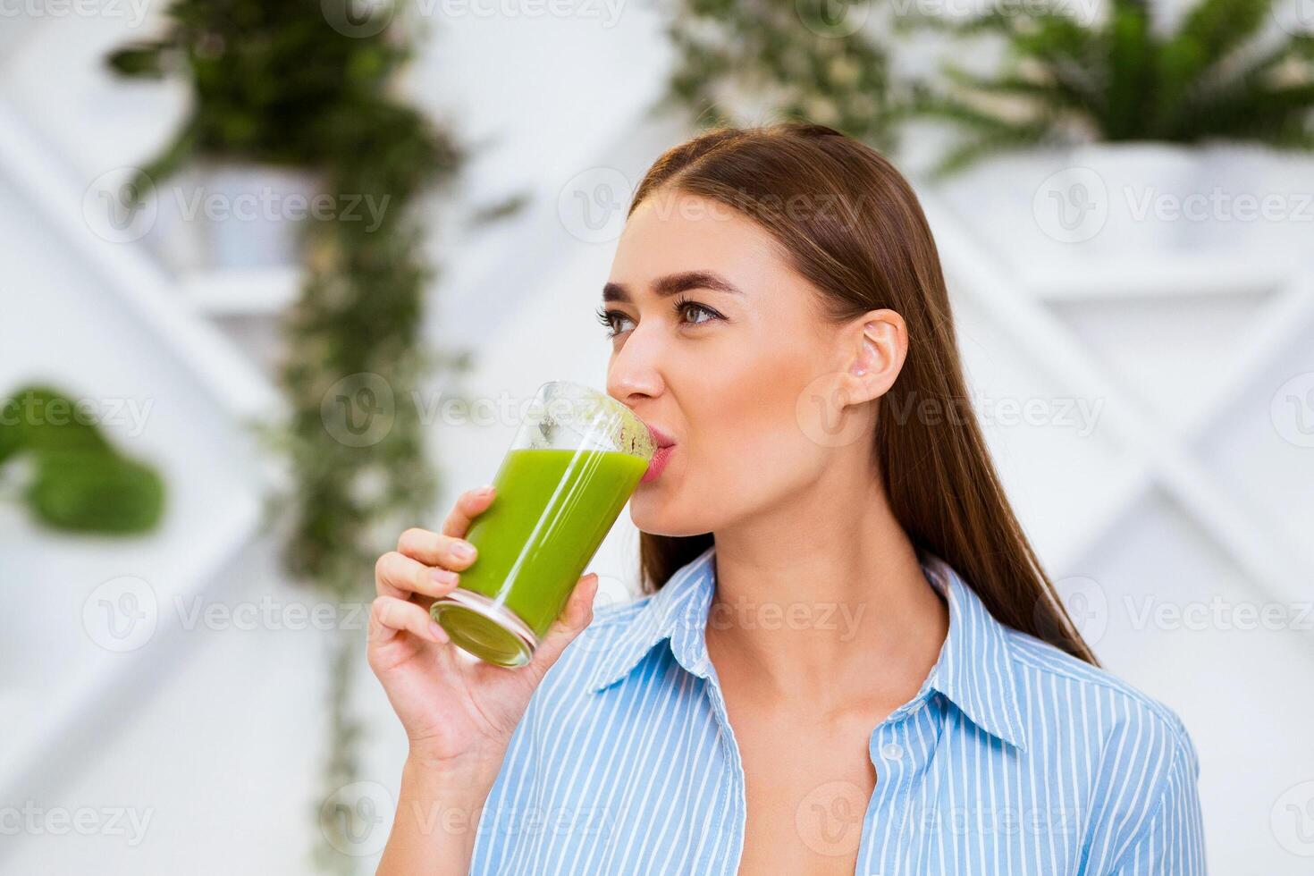 A woman wearing a blue striped shirt drinks from a glass of green smoothie. Shes standing in a bright, modern setting with plants in the background. photo