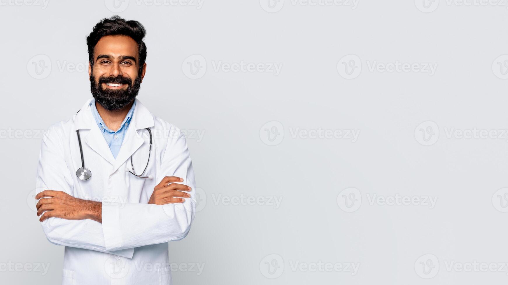 Indian man doctor with a dark beard and a white lab coat stands against a white background with his arms crossed and smiling at the camera. He is wearing a stethoscope around his neck, copy space photo